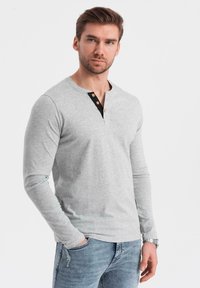 Grijze langemouwenshirt met een henley-halslijn, voorzien van een zwarte knoopsluiting en bruine knopen. Draagt met denim broek en een horloge.