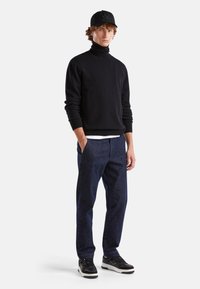 Maglione nero a collo alto, pantaloni di jeans blu scuro e sneakers nere con dettagli bianchi. Indossato con un berretto nero con un logo.