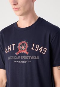 Tricou din bumbac de culoare bleumarin, cu decolteu rotund, având un design grafic cu un logo care include "1949" și "AMERICAN SPORTSWEAR" în text roșu și alb.