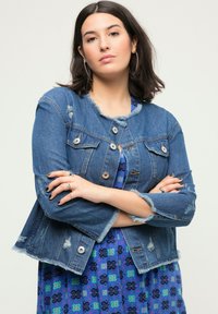 Chaqueta de denim con cuello deshilachado, detalles desgastados y botones de metal. Lleva puesta sobre un vestido azul con estampado y acentos en verde y púrpura.