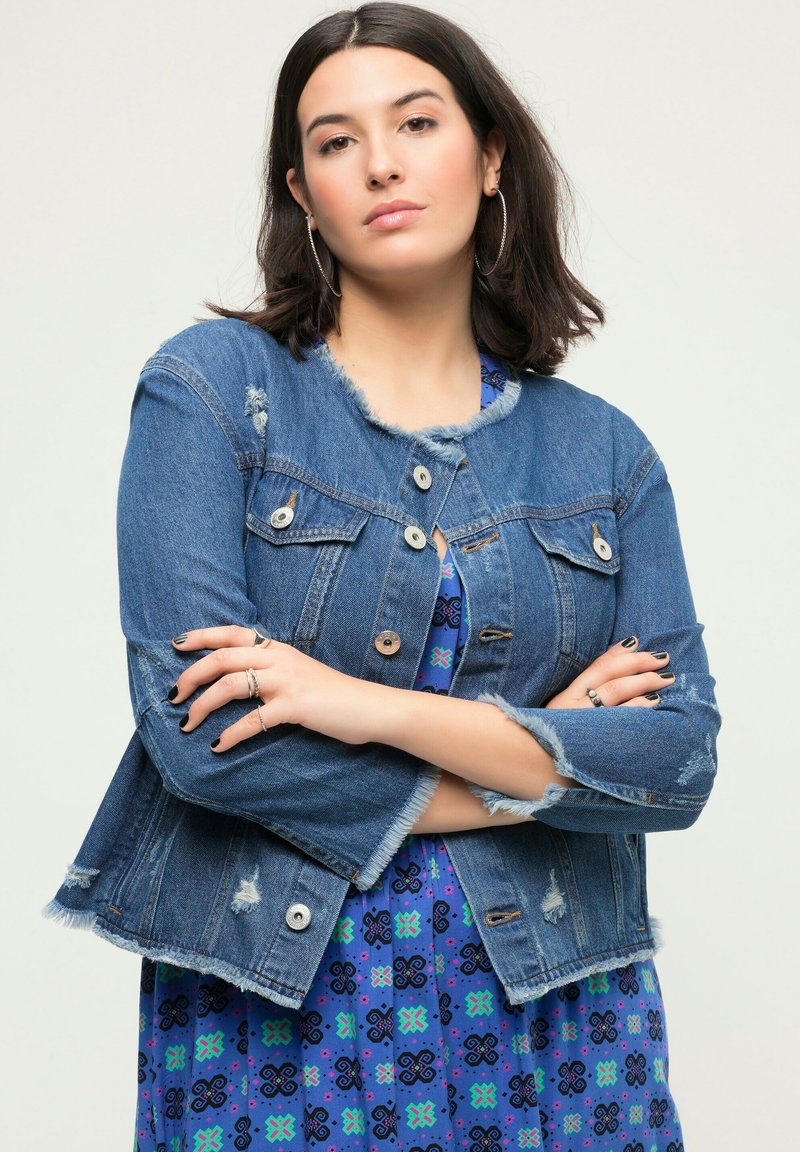 Chaqueta de denim con cuello deshilachado, detalles desgastados y botones de metal. Lleva puesta sobre un vestido azul con estampado y acentos en verde y púrpura.