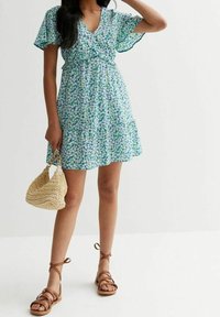 Vihreä-sininen kukallinen lyhyt mekko, kevyt kangas, röyhelöiset hihat, istuva vyötärö, kerroksellinen hame. Käsinkehrätty laukku ja ruskeat remmilenkkarit.