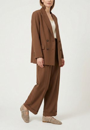 Frau trägt braunen zweireihigen Blazer und weitgeschnittene Hose mit beigefarbenen perforierten Mary-Jane-Schuhen, steht vor einfachem Hintergrund.