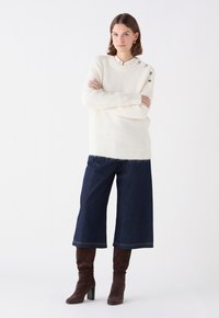 Maglione bianco lavorato a maglia con vestibilità rilassata, caratterizzato da tre bottoni decorativi sulla spalla, abbinato a jeans blu scuro a gamba larga e stivali marroni al ginocchio.