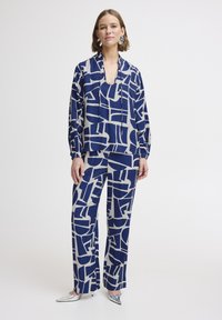 Blusa a fantasia navy e beige con pantaloni a gamba larga coordinati in un tessuto morbido. Caratterizzata da forme geometriche e una vestibilità rilassata. Scarpe argentate in evidenza.