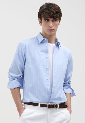 Mann in hellblauem, gestreiftem Hemd mit hochgekrempelten Ärmeln, weißem T-Shirt, weißen Hosen und braunem Gürtel, die Hände in den Taschen, schauend nach rechts.