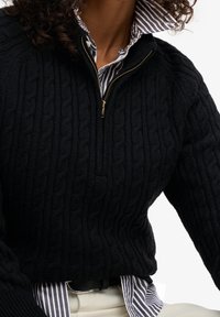 Personne portant un pull noir en maille torsadée avec fermeture éclair par-dessus une chemise rayée bleue et blanche boutonnée, un pantalon beige et une ceinture noire.