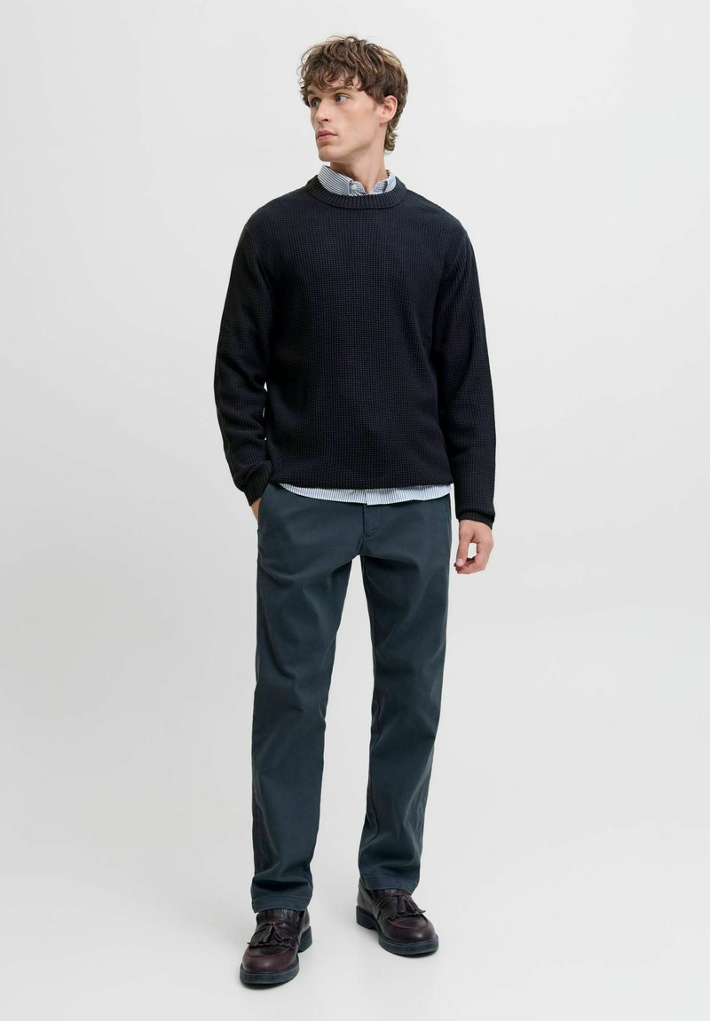 Suéter negro de punto con cuello redondo, superpuesto a una camisa de rayas azul claro, combinado con pantalones gris oscuro y zapatos marrones.