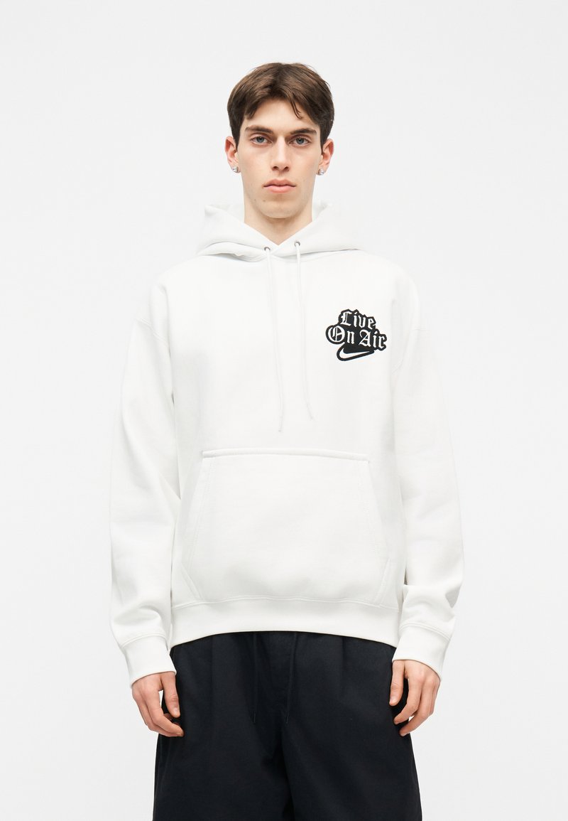 Junger Mann trägt einen weißen Hoodie mit dem Aufdruck "Live On Air" und Logo auf der Brust, kombiniert mit lockeren schwarzen Hosen, und steht vor einem schlichten Hintergrund.