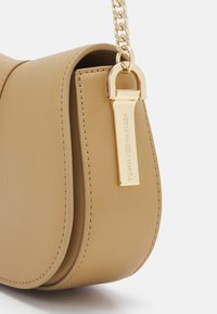Borsa a tracolla in pelle color tan con design curvo, tracolla in catena dorata e un ciondolo rettangolare con dettagli del logo in rilievo.