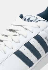 Λευκό sneaker από δέρμα με τρεις ναυτικές ρίγες, υφή με λεπτομέρειες, λευκά κορδόνια και ναυτικό εσωτερικό πάτο· το λογότυπο στη γλώσσα έχει σχέδιο με τρία φύλλα.