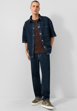 Homme debout contre un mur portant une chemise en denim bleu foncé à manches courtes, un jean en denim assorti, un t-shirt marron et des baskets beiges.