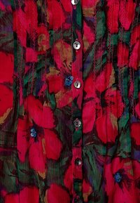 Textilblus med ett livligt blommönster i röd, grön och lila. Inkluderar stora knappar och små blå blommor som dekoration.