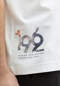 Hvid bomulds T-shirt med et grafisk design med "1962" og teksten "HVOR ØDELAND MØDER HAVET" i sort og flerfarvede accenter.
