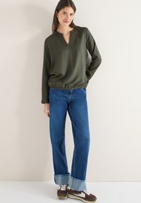 Blouse verte à manches longues avec un col en V et un bas froncé, associée à un jean bleu ample avec des ourlets effilochés et des baskets marron.