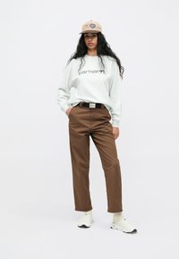 Szara bluza z logo "Carhartt", brązowe spodnie, czarny pasek, beżowa czapka i białe buty z czarnymi akcentami. Prosty design i luźny krój.