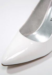 Chaussure à talon haut blanche en cuir verni à bout pointu, doublure intérieure argentée, vue de face supérieure sur un fond blanc.