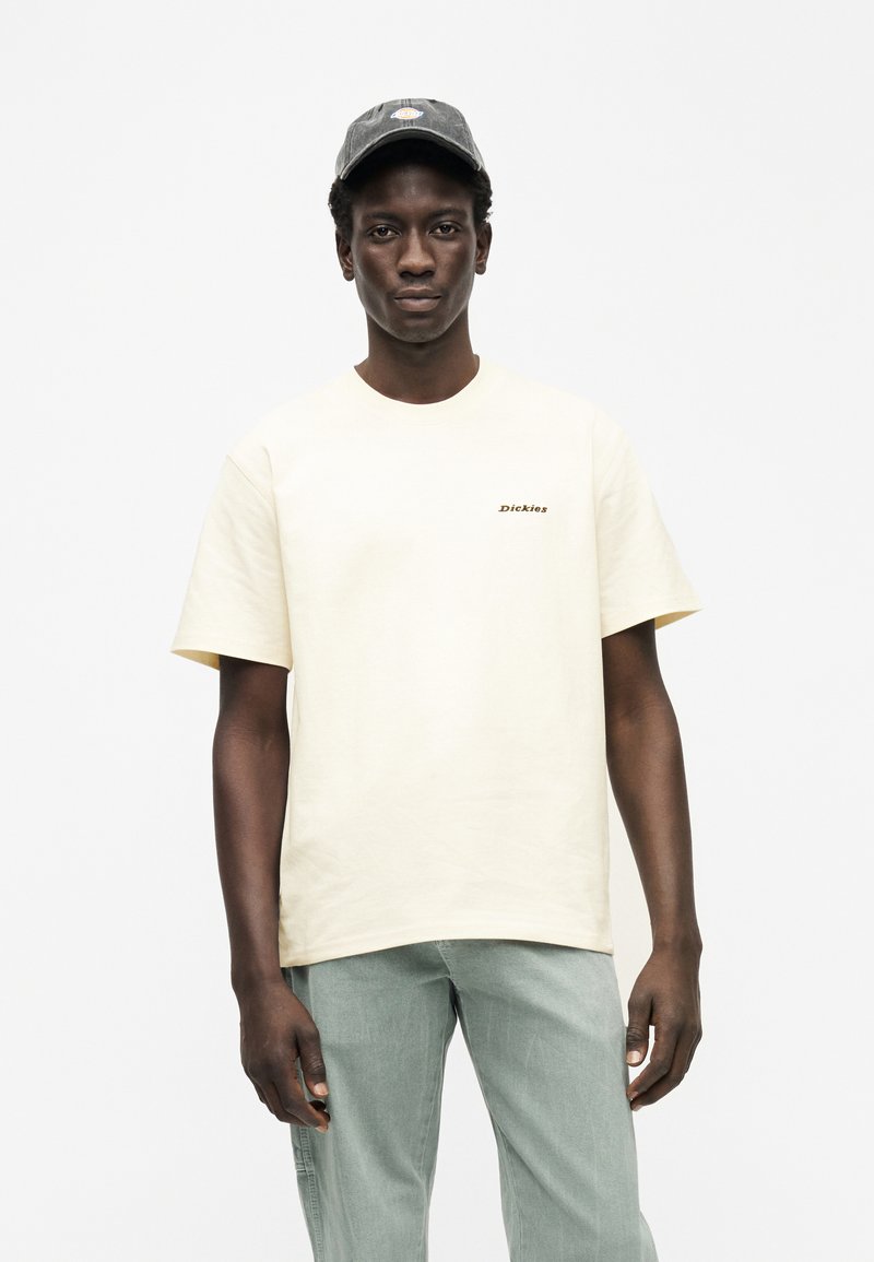 T-shirt in cotone color crema con maniche corte e scollo rotondo, con un piccolo logo "Dickies" marrone sul petto. Pantaloni verde chiaro.