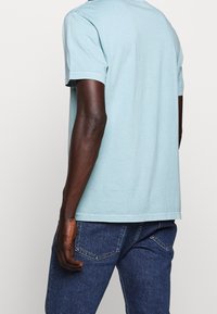 T-shirt bleu clair à manches courtes avec un col rond, en coton doux. Présente une coupe droite et des détails de couture minimaux. Porté avec un jean foncé.