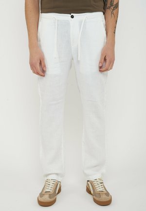 Man draagt witte linnen broek met trekkoord, bruine en beige sneakers en een donker overhemd, staand tegen een effen lichte achtergrond.