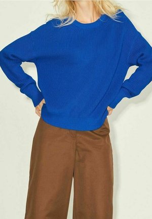 Personne portant un pull tricoté bleu vif à manches longues et un pantalon marron ample, les mains posées sur les hanches sur un fond uni.