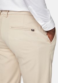 Bežās bikses ar faktūrētu audumu, ar aizmugurējo kabatu, kurā atrodas neliels logotipa uzlīme. Kopā ar baltu pogu krekli.