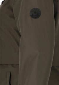 Olijfgroene waterdichte jas met een gladde textuur, voorzien van een ronde zwarte logopatch op de mouw en ritszakken.