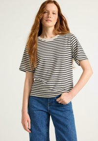 Gestreiftes T-Shirt in Beige und Schwarz, aus weichem Stoff, mit weit geschnittenen kurzen Ärmeln und bequemer Passform. Kombiniert mit blauen Jeans.
