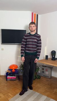 Striped sweater with green, burgundy, and white colors; black straight-leg jeans; dark polished shoes; tattoos visible on hands.