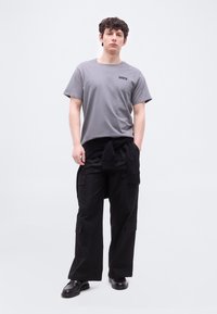 Νέος άνδρας που στέκεται, φορώντας γκρι μπλουζάκι Levi's, μαύρο παντελόνι cargo, μαύρο μπουφάν δεμένο γύρω από τη μέση και μαύρα δερμάτινα παπούτσια σε λευκό φόντο.