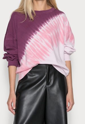 Kvinde iført en langærmet tie-dye sweatshirt i lilla, lyserød og hvid med sorte læderbukser, vist fra nakken til midten af låret.