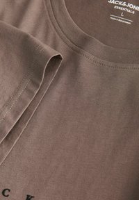 Brun bomulds T-shirt med rund halsudskæring, korte ærmer og synlige sømme. Har sort tekst nær kanten og labels for størrelse og oprindelse.