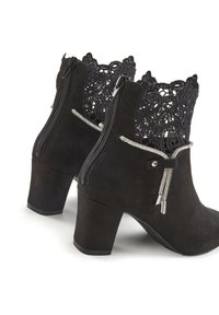 Botas de tobillo de ante negro con una textura en forma de encaje en la parte superior, cremalleras laterales y detalles decorativos en plata en las cremalleras y el contorno.