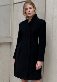 Czarny wełniany płaszcz o strukturalnym wzornictwie, z długimi rękawami, wysokim kołnierzem i dopasowanymi szwami. Gładka tekstura z subtelnym połyskiem, do kolan.
