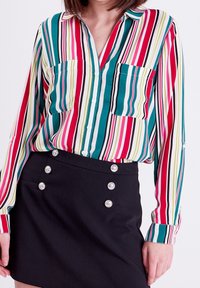 Chemise à boutons rayée en bleu sarcelle, rose et vert ; comporte deux poches poitrine et une coupe décontractée. Assortie à une mini-jupe noire avec des détails à boutons.