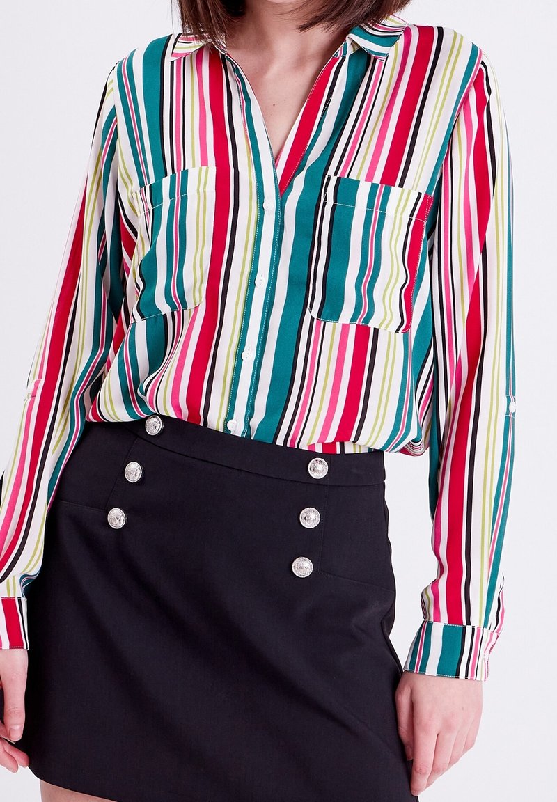 Chemise à boutons rayée en bleu sarcelle, rose et vert ; comporte deux poches poitrine et une coupe décontractée. Assortie à une mini-jupe noire avec des détails à boutons.