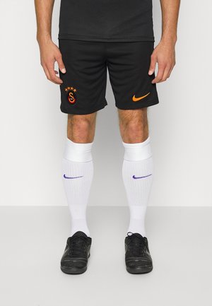 Mężczyzna w czarnych szortach piłkarskich Galatasaray, białych skarpetach z fioletowymi logo Nike oraz czarnych butach sportowych, stojący na jednolitym tle.