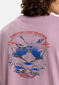 Lyslilla bomulds T-shirt med et grafisk print af en vulkan, palmer og teksten "TASTES LIKE HEAVEN" i fed rød og blå farve.