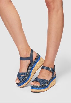 Jambes portant des sandales compensées tissées bleues avec des brides cheville et une semelle plateforme multicolore en fibres naturelles sur un fond uni.