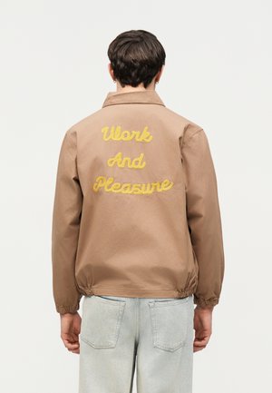 Persoon die een beige jas draagt met gele geborduurde tekst "Work And Pleasure" op de rug, gecombineerd met lichtblauwe spijkerbroek.