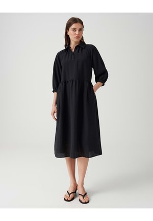 Vrouw draagt een zwarte midi-jurk met lange mouwen en knopen, met ingezette taille en zakken, gecombineerd met zwarte sandalen, staand tegen een effen achtergrond.