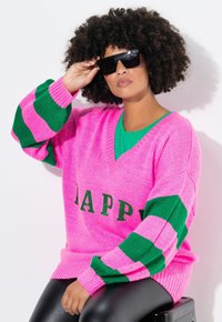Roze trui met groene gestreepte mouwen en "HAPPY" in groene letters. Gelaagd over een groene top, gecombineerd met zwarte leggings en een zonnebril.