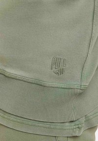 Tissu de sweatshirt vert clair avec bord côtelé, arborant un logo embossé "PULL IN". Le matériau est lisse avec une texture subtile.