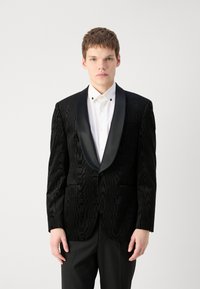 Svart tuxedojacka med texturerad tyg, satinreversar, en ficka, två framfickor, bärs över en vit skjorta med knappar.