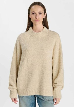Vrouw draagt een losse, beige fuzzy trui met lange mouwen en blauwe spijkerbroek, staand tegen een effen witte achtergrond.