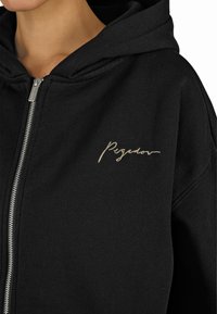 Fekete, cipzáras kapucnis pulóver, puha anyagból készült, ezüst cipzárral. A bal mellen egy arany írással készült "Pegador" logó található.