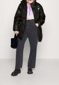 Manteau en duvet noir, pull graphique rose, jeans larges gris foncé, chaussures à plateforme noires, tenant un sac en fourrure noir. Tenue moderne et décontractée.