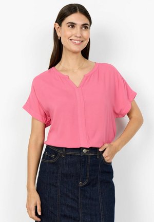 Uśmiechnięta kobieta w różowej bluzce z krótkim rękawem i ciemnej dżinsowej spódnicy, z jedną ręką w kieszeni, na jednolitym białym tle.