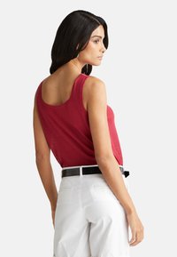 Top senza maniche rosso, realizzato in materiale leggero, con una vestibilità rilassata e schiena arrotondata; abbinato a pantaloni corti bianchi e una cintura nera.