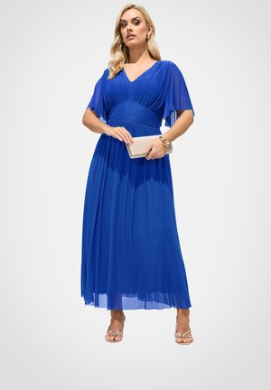 Frau trägt ein fließendes, königsblaues V-Ausschnitt-Kleid, hält eine beige Clutch, dazu goldene Sandalen und Armbänder, steht vor einem schlichten Hintergrund.